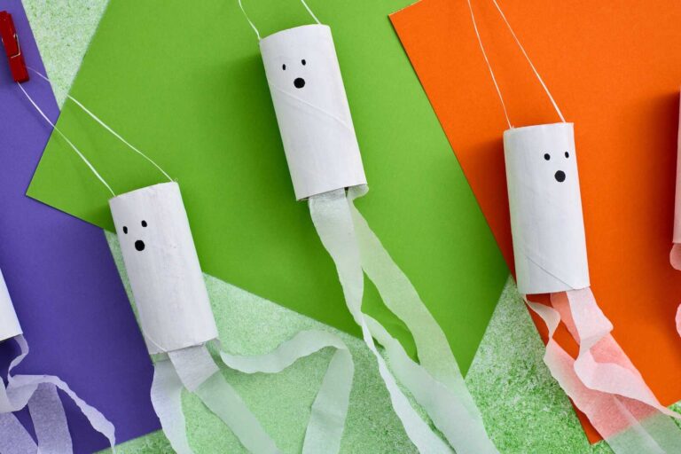 Several toilet-paper-roll ghost crafts hang on white string, arranged over overlapping sheets of bright purple, green, and orange paper. Each roll is painted white with a simple black ghost face, and long white tissue-paper streamers trail from the bottom, creating a floating, playful Halloween look.