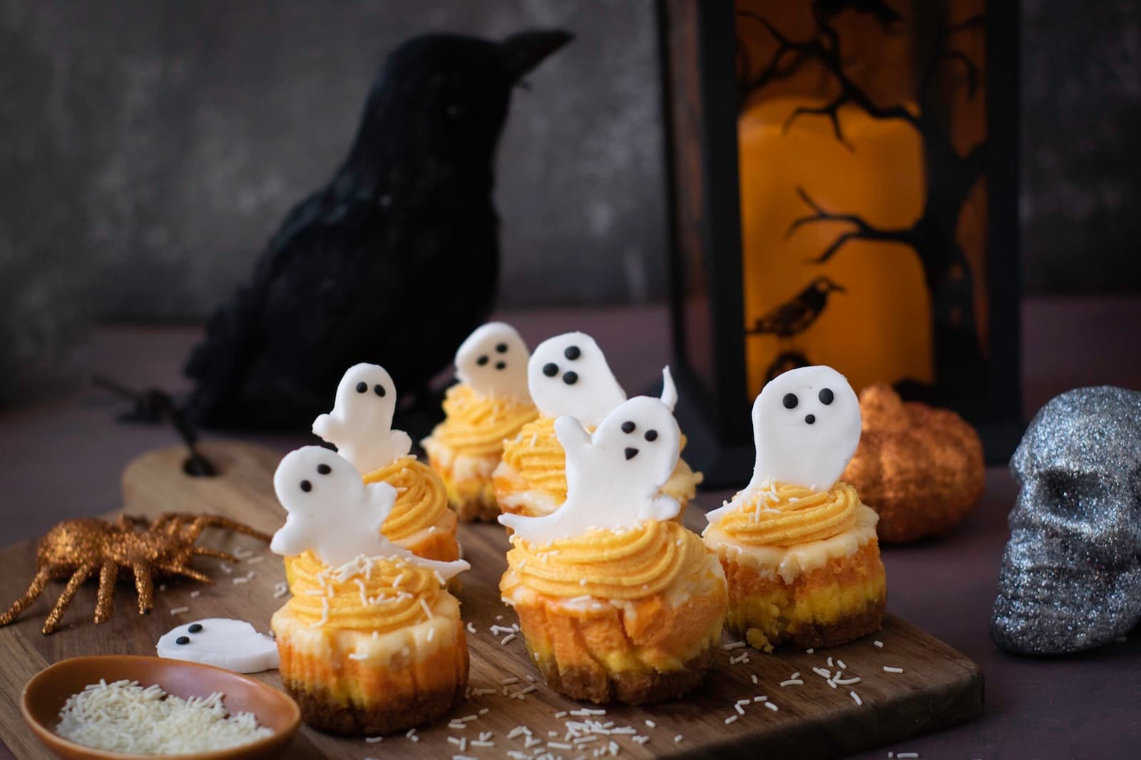 Halloween Candy Corn Mini Cheesecakes topped with ghost decorations on a wooden board with spooky Halloween decor.