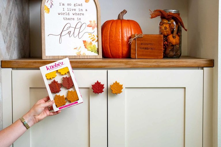 Hand holding a 6 pack of Fall Leaf Knobēz beside red and yellow leaf knobs on cream cabinets with fall decor.